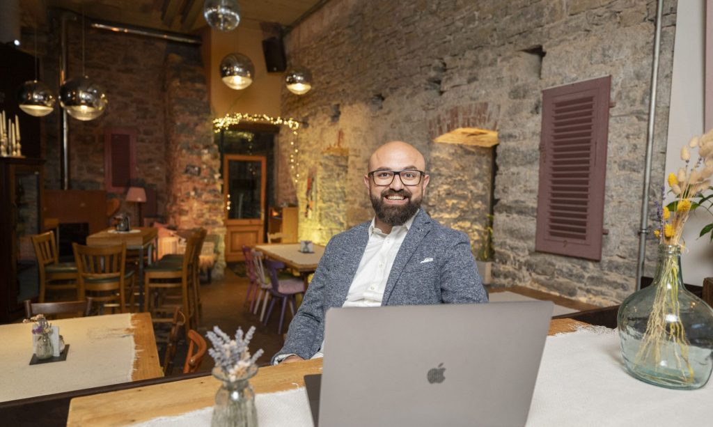 Gökhan sitzt an einem Tresen mit dem aufgeklappten Laptop und lächelt seinem neuen Begleiter entgegen